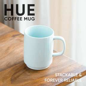 White coffee mug on a wooden surface with 'HUE' branding and text about stackability and reliability.