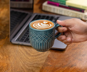 Person holding a teal mug with a latte art design in front of a laptop on a wooden desk.