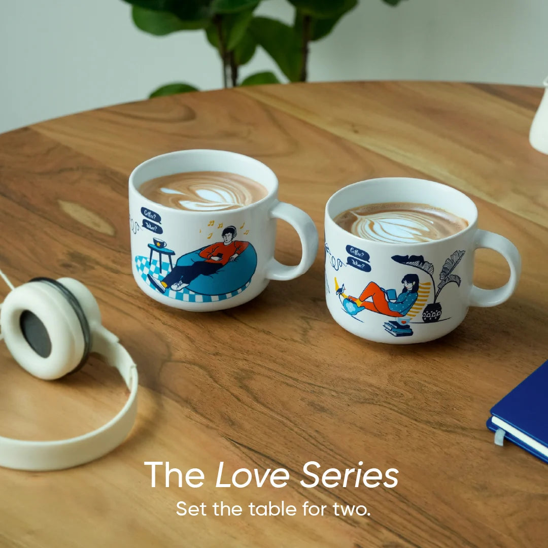Two mugs with artistic designs on a wooden table, accompanied by headphones and a notebook.