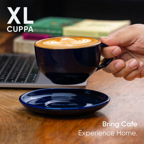 Hand holding a black coffee cup with latte art on a wooden table with books and a laptop.
