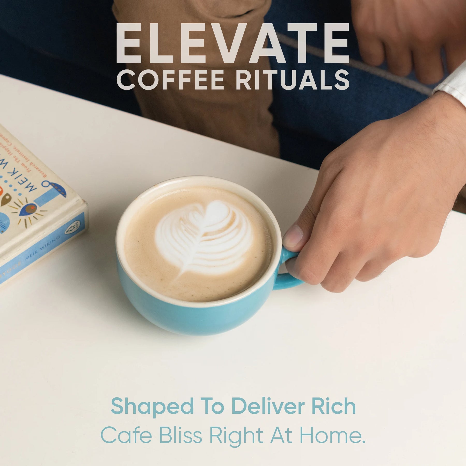 Person holding a blue mug with a cappuccino, next to a book, on a white surface with text 'Elevate Coffee Rituals'.